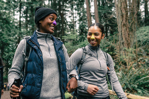 Two women hiking in nature surrounded by trees while wearing Nöz colorful zinc oxide sunscreen formula for the nose.